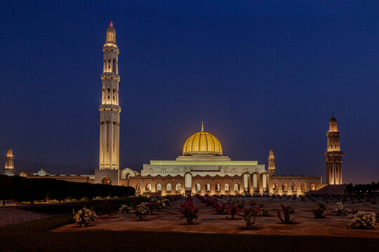 View of the illuminated Sultan Qaboos Grand Mosque at night with its golden dome and minarets rising above the manicured gardens in Muscat, Muscat Governorate, Oman.