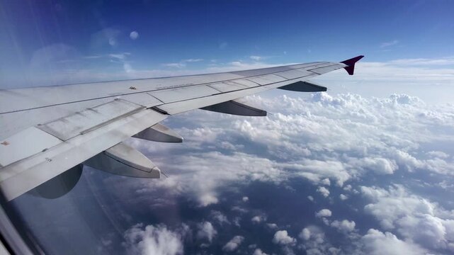Scenic passenger POV aerial view from an airplane window, showcasing the aircraft wing soaring high above a beautiful sea of fluffy white cumulus clouds under a clear blue sky during a daytime flight 