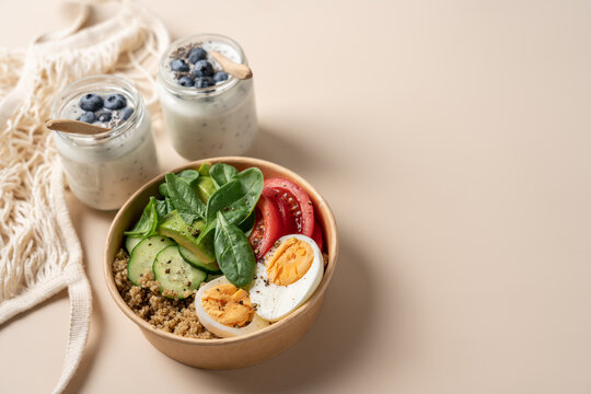 Healthy protein bowls with  egg, quinoa, avocado, spinach, cucumber  in cardboard containers, served with chia seed yogurt and blueberries in glass jars on a light background, balanced nutrition