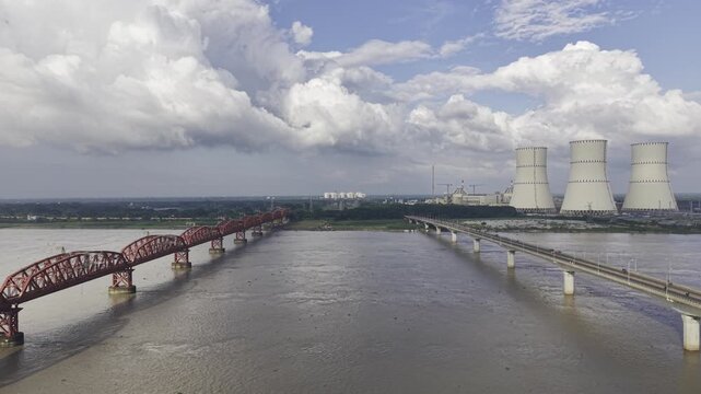 Backward drone footage between Hardinge Bridge and Lalon Shah Bridge over the Padma River, capturing Rooppur Nuclear Power Plant cooling towers, Green City Rooppur buildings, blue sky and clouds.
