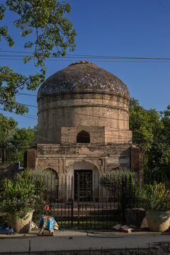 View of the historic 17th-century Mughal-era Buddhu ka Awa tomb featuring an intricate tiled dome and weathered brick walls behind a metal fence in Lahore, Punjab, Pakistan.