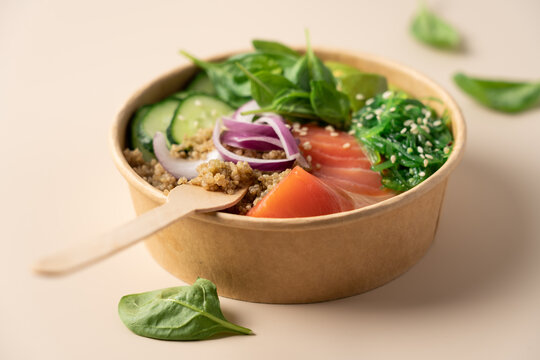 Healthy protein bowl with quinoa, smoked salmon, spinach, cucumber, avocado and chuka seaweed salad in a cardboard container on a light beige background, balanced nutritious meal 