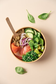 Healthy protein bowl with quinoa, smoked salmon, spinach, cucumber, avocado and chuka seaweed salad in a cardboard container on a light beige background, balanced nutritious meal 