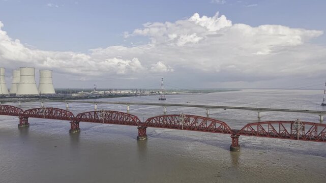 Aerial view of the Padma River between Hardinge Bridge and Lalon Shah Bridge, featuring Rooppur Nuclear Power Plant cooling towers, Green City Rooppur buildings, blue sky and scattered clouds.