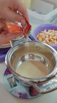 High-angle close-up shows hands squeezing orange seasoning packet over stainless steel pot resting on digital kitchen scale while thin stream falls during careful food prep and portion control