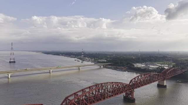 Diagonal forward drone footage approaching Hardinge Bridge and Lalon Shah Bridge over the wide Padma River, showing sections of both bridges, riverbanks and dramatic clouds across the sky.