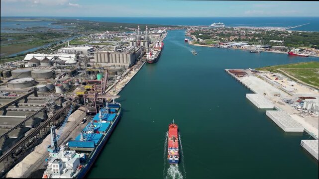 Top down aerial drone view of cargo ship sailing along Ravenna port canal past docked vessel and industrial grain silos in Emilia Romagna, Italy