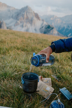 View of a person pouring water from a bottle into a camping pot on a portable stove in a grassy mountain field with Piz da Peres in the background in Mareo, Trentino-Alto AdigeS, Italy.