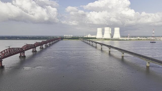 A slow descending aerial view over the Padma River between Hardinge Bridge and Lalon Shah Bridge, with Rooppur Nuclear Power Plant cooling towers, Green City Rooppur buildings, blue sky, and clouds.