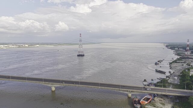 Aerial ascending drone footage of Lalon Shah Bridge crossing the Padma River, with Rooppur Nuclear Power Plant cooling towers, cargo boats, blue sky, and white clouds visible in Bangladesh.
