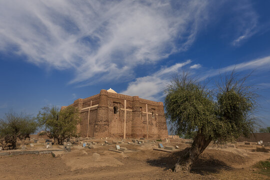 View of the historic Khalid Walid Tomb with its distinctive brick architecture set within a cemetery under a bright blue sky in Kabirwala, Punjab, Pakistan.