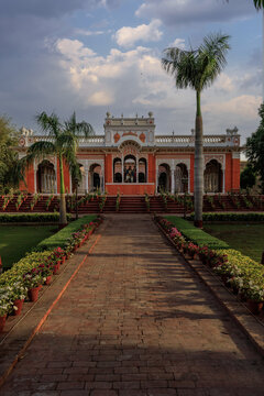 View of the Bara Dari at Darbar Mahal with its distinctive red facade and arched architecture, framed by palm trees and a brick pathway lined with potted plants in Bahawalpur, Punjab, Pakistan.