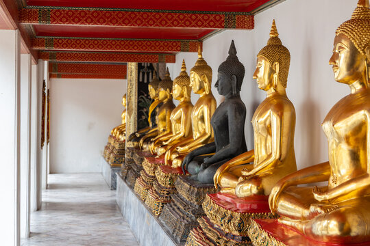 Das Bild zeigt eine beeindruckende Reihe vergoldeter Buddha-Statuen sowie eine markante dunkle Statue in einer Wandelhalle des Wat Pho in Bangkok, Thailand