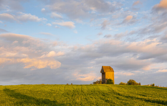 View of Wiatrak Kozlak Jan, a historic wooden windmill, standing in a lush green field under a soft sunset sky in Suchynicze, Podlaskie Voivodeship, Poland.