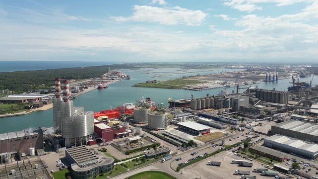 Aerial drone view of red gas tanker ship docked at Ravenna port near chemical refinery plant with gas flare stack burning bright on sunny day in Emilia Romagna Italy 