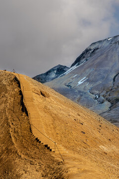 View of the hiking trail with stairs winding up the golden-brown volcanic slopes of Kerlingarfjoll with snow-capped peaks in the background, Hrunamannahreppur, Iceland.