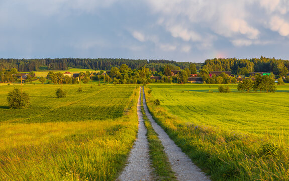 View of a dirt road leading through green fields towards a village and forest under a dramatic golden hour sky in Sokolka, Podlaskie Voivodeship, Poland.