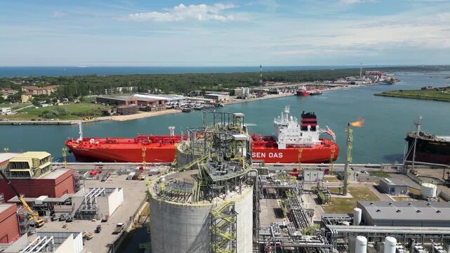 Aerial drone view of red gas tanker ship docked at Ravenna port near chemical refinery plant with gas flare stack burning bright on sunny day in Emilia Romagna Italy 