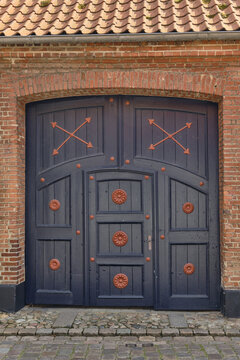 View of a traditional dark blue wooden door with red iron decorative accents and studs set into a historic red brick wall on a cobblestone street in Ribe, Denmark.