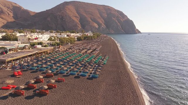 Drone view along Perissa black sand beach with rows of straw umbrellas towards Mesa Vouno mountain Santorini
