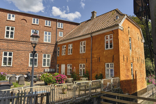 View of a historic orange stucco building and an adjacent red brick structure with white window frames and a small outdoor seating area with flowers in Ribe, Denmark.