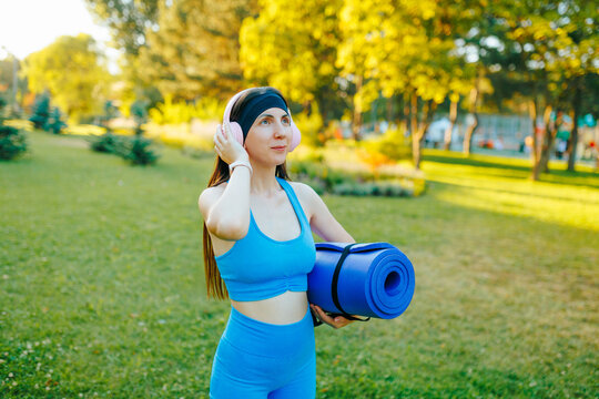 Active fit woman holding a yoga mat and adjusting headphones in the park.