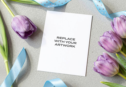 Vertical stationery mockup featuring a blank white card on textured surface surrounded by purple tulips and light blue ribbons, floral arrangement with gift wrapping elements, Template