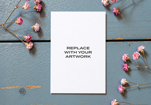 Vertical paper mockup with replace your artwork text displayed on a blue textured wooden surface surrounded by pink dried flowers, stationery branding presentation, print design template