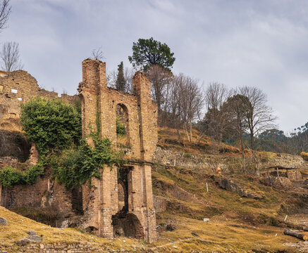 View of the historic ruins of the Murree Brewery with weathered brick walls, arched window frames, and climbing vines set against a hillside landscape in Murree, Punjab, Pakistan.
