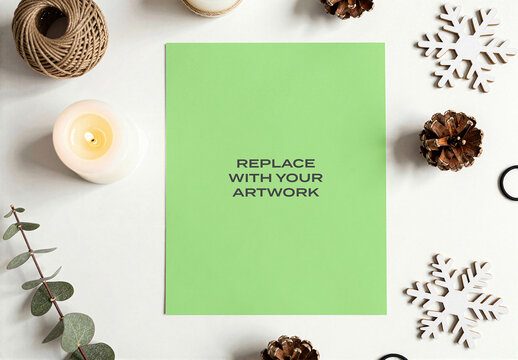 Poster mockup displayed on a white surface in flat lay stationery presentation with pinecones, eucalyptus, candles, twine, and snowflake ornaments, natural lighting, Template
