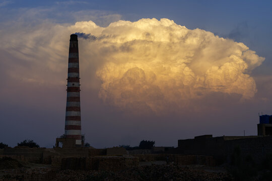 View of a tall striped brick kiln chimney emitting smoke against a dramatic golden cloud formation at sunset in Harappa, Punjab, Pakistan.