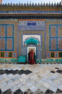 View of the intricate tile work at the entrance of Darbar Hazrat Sultan Bahoo, with a woman in a red veil and children standing on the marble floor in Basti Samundri, Punjab, Pakistan.