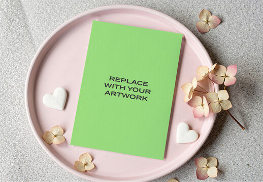 Vertical card mockup on a pink ceramic plate with dried hydrangea flowers and white heart-shaped candies, flat lay stationery presentation textured surface natural light, print design template
