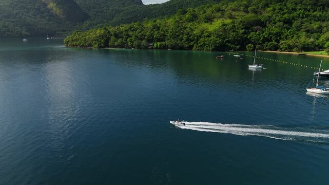 Motor boat moves fast on the calm and green lagoon with anchored sailing boats on the island of Ilha Grande, Brazil