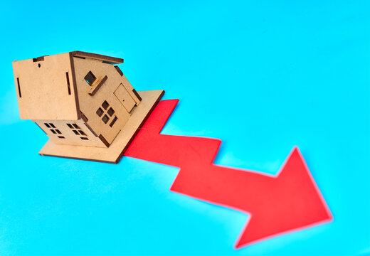 Wooden house model with large red falling arrow showing real estate market decline on turquoise background
