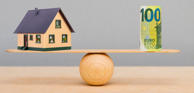 Auf einer Wippe ist ein Modellhaus und die symbolische Finanzierung im Gleichgewicht - FOTOMONTAGE