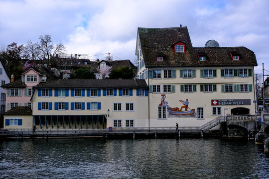 Zurich, Switzerland - March 15, 2026. A serene waterfront view of traditional Swiss architecture in the historic Schipfe artisan district, featuring building facades with distinctive blue shutters