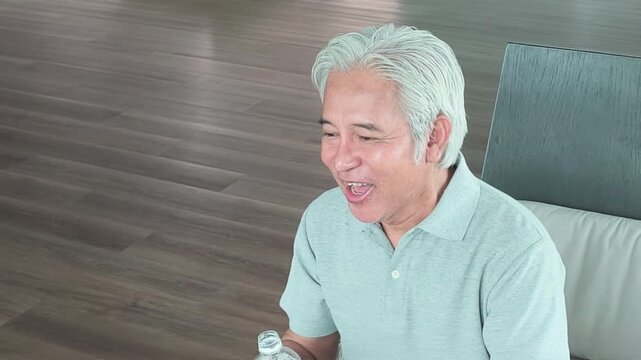 asian senior man sitting and drinking water from a plastic bottle in his living room, feeling refreshed and hydrated