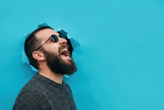 Happy Bearded Man with Sunglasses Laughing and Breaking Through a Blue Paper Background with Copy Space