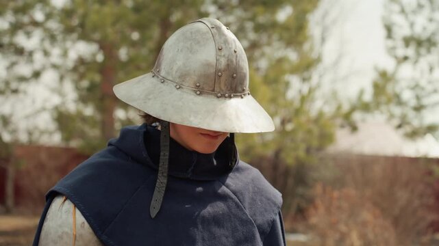 Reenactor in historical armor lifts helmet to reveal face and chin, showing clear upward motion and visible change from concealed to open expression in warm daylight outdoors, with medieval