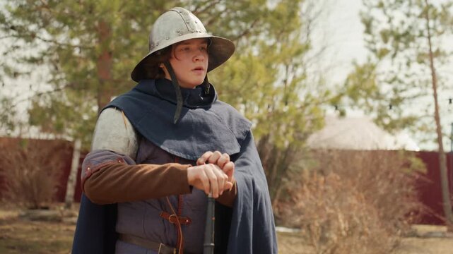 Young person wearing helmet, padded gambeson, cloak, and shoulder armor stands outdoors in rustic village yard, gripping sword hilt while turning head to survey surroundings in calm daylight