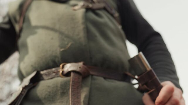 Low angle scene showing hands gripping sword hilt and scabbard while blade is drawn at waist, dark green tunic, black sleeves, leather belt, and focused outdoor light, with sword, unsheathing