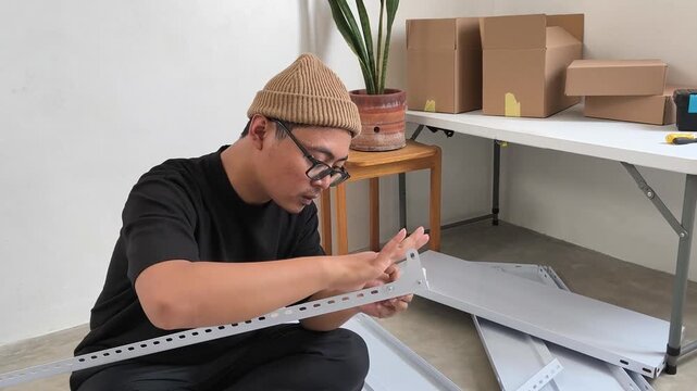 Asian man assembling white metal storage shelf frame at home, furniture assembly, storage rack installation, room organization, handyman work, and self-installation.