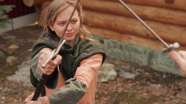 Young woman performs controlled sword techniques outdoors in rustic rustic setting, moving from steady guard into forward thrust and body turn while keeping focused, confident expression and
