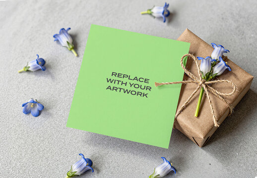 Square card mockup with text placeholder positioned next to a gift box wrapped in brown paper and tied natural twine blue flowers on textured gray surface, Template