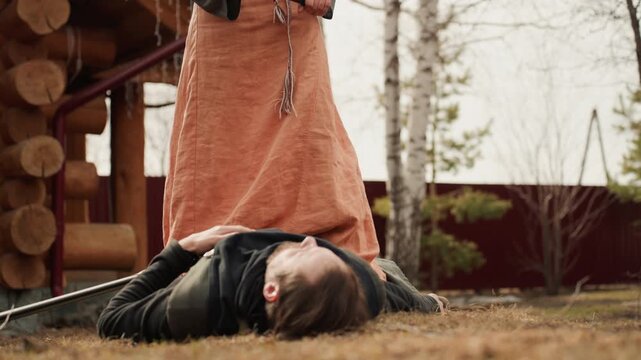 Young woman in medieval leather and cloth costume lies prone on dry grass beside wooden cabin while standing companion with sword and long dress remains visible in dramatic outdoor historical setting