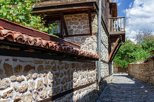 A narrow cobblestone street winds between traditional stone houses with wooden balconies and tiled roofs under a blue sky with clouds.
