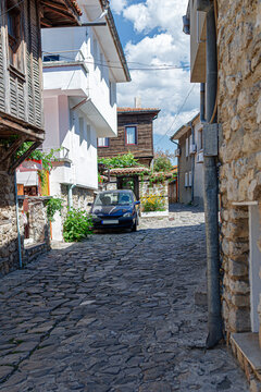 A dark car is parked on a narrow cobblestone street surrounded by traditional stone and wooden houses under a sunny sky.