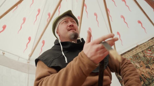Middle-aged man in medieval costume grips wooden staff and speaks inside patterned canvas tent, with expressive hand movement, soft daylight, and candid historical performance mood