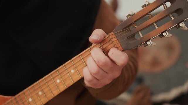 Adult hands on acoustic guitar neck near headstock with fingers pressing strings and thumb wrapped over neck to form chord, showing controlled grip, focused fingering, and calm playing posture indoors